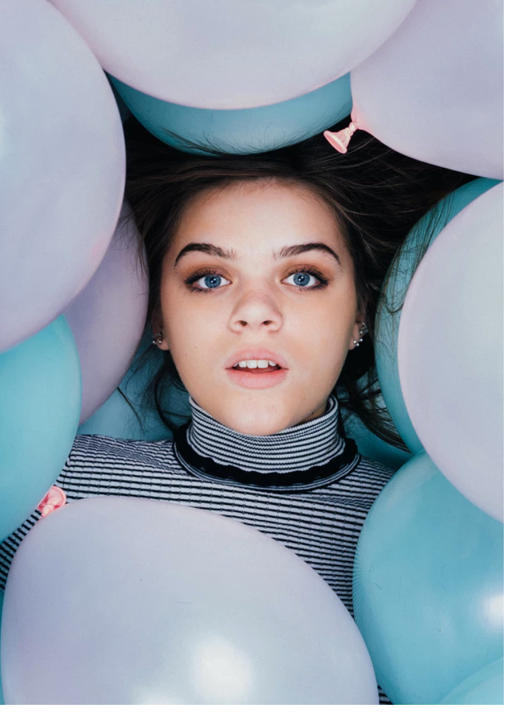 A photo of Harper starring straight into the camera surrounded by teal and pink balloons. Her hair is dark brown, her eyes are steel blue, and her mouth is slightly agape.