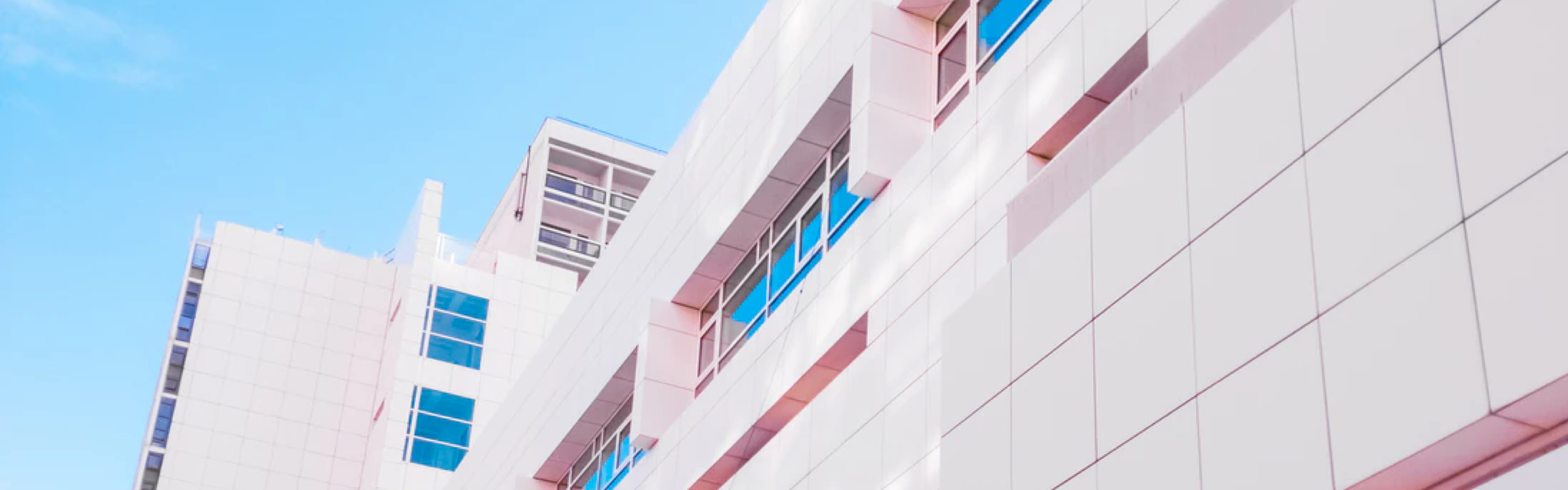 A dutch angle looking up at the corners of two white apartment buildings against a clear blue sky. A subtle pink filter has been applied to the image.