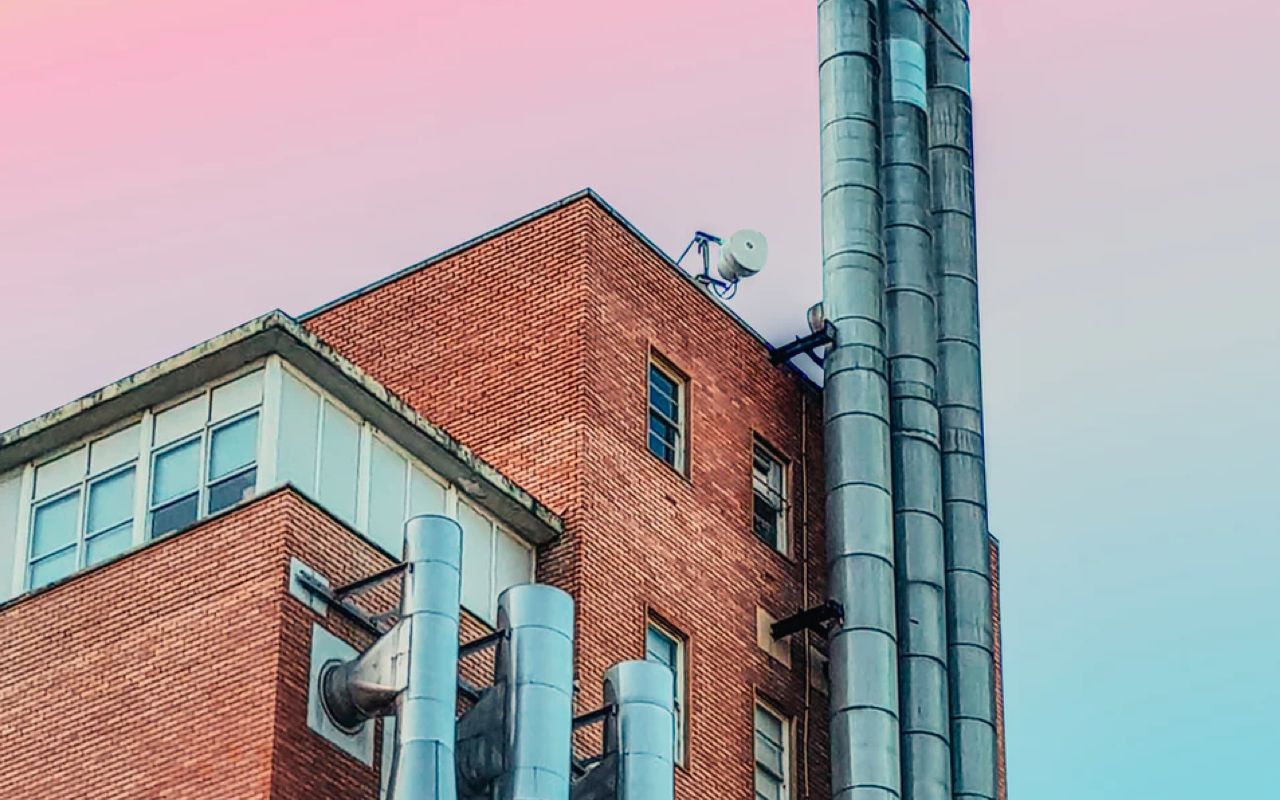 An image of a red brick factory building with several tall steel ventilation pipes lining the side of the building. The sky behind the building is a faded pink.