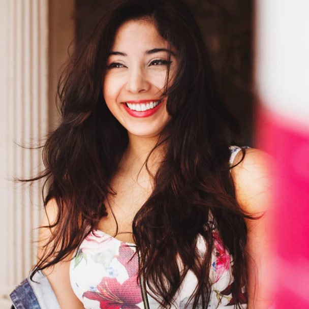 A photo of Harper smiling brightly. Her long hair is down over her shoulders and she is wearing a white sundress with pink flower patterns.